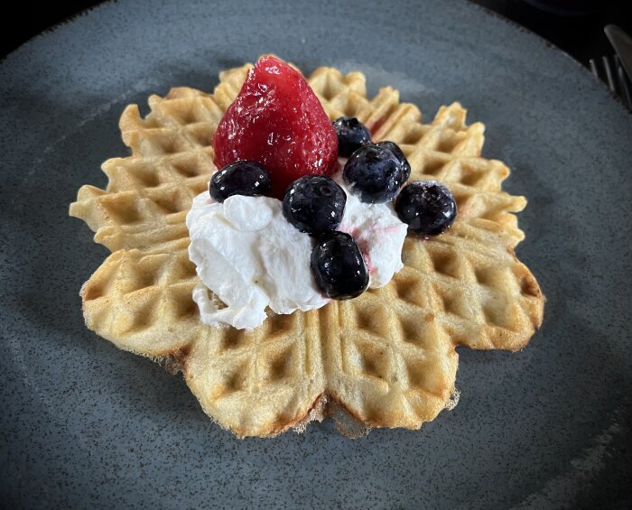 Våffla med grädde, jordgubbar, blåbär på mörk tallrik.