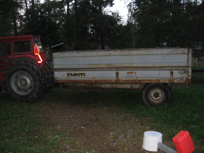 Röd traktor kopplad till en sliten grå släpvagn med texten "TUHTI", omgiven av träd i skymningen.