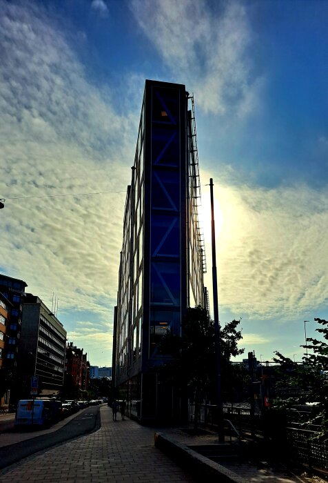 Modern byggnad med glasfasad mot en dramatisk himmel, stadsvy, gator, några gående personer och parkerade bilar.