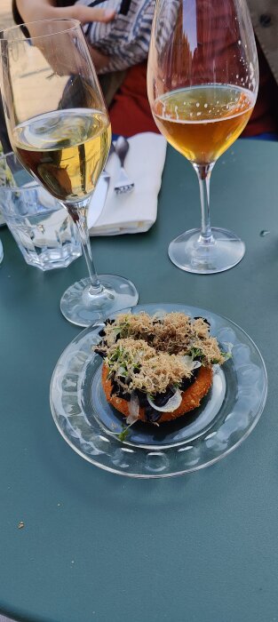 Gourmetmat på tallrik, vin och öl i glas, personer vid restaurangbord utomhus.