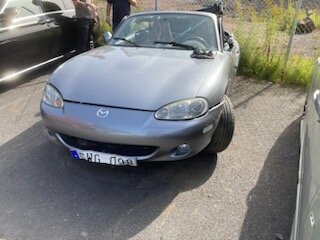 Grå Mazda Miata med framhjulspunktering parkerad vid sidan av vägen, soligt väder, andra fordon i närheten.