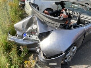 Bakdel av en silverfärgad cabrioletbil skadad i kollision, parkerad vid sidan av vägen med vegetation.