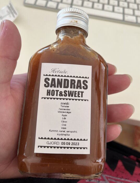 En hand håller en flaska märkt "SANDRAS HOT&SWEET" med ingredienser som tomat och chili. Tillverkningsdatum: 05/09/2023.