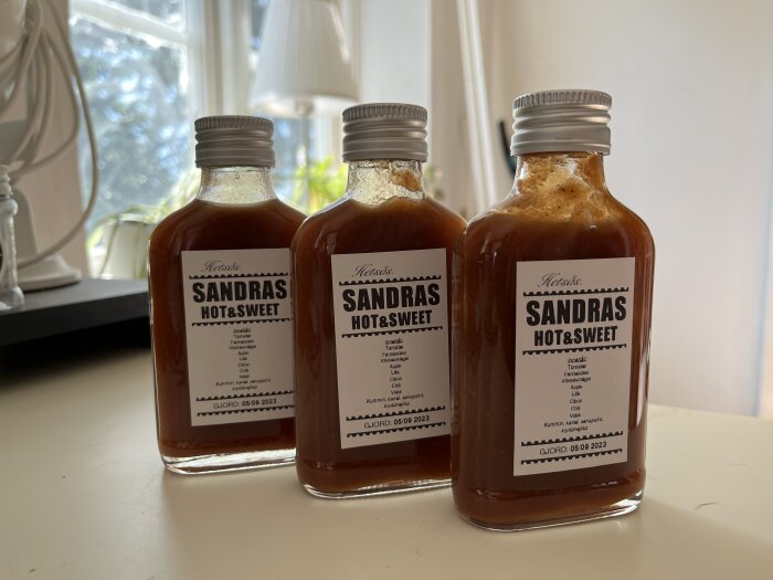 Tre flaskor med märket "SANDRAS HOT&SWEET" står på fönsterbräda; sannolikt kryddig sås.