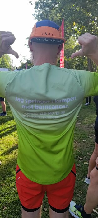 Person i träningskläder, keps bakochfram, visar muskler, text mot barncancer på t-shirt.