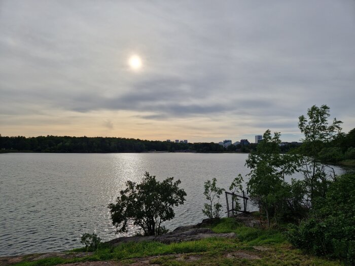 Molnigt himmel, solreflektion på vattnet, stadsilhuett i bakgrunden, gröna träd, klippig strandkant.