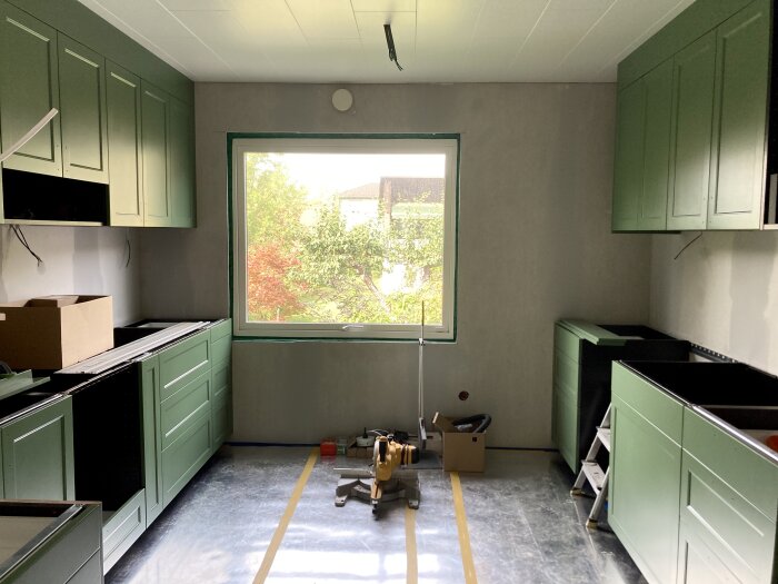 Kök under renovering, grön skåpsinredning, kartonger, byggverktyg, golvskydd, stort fönster med utsikt.