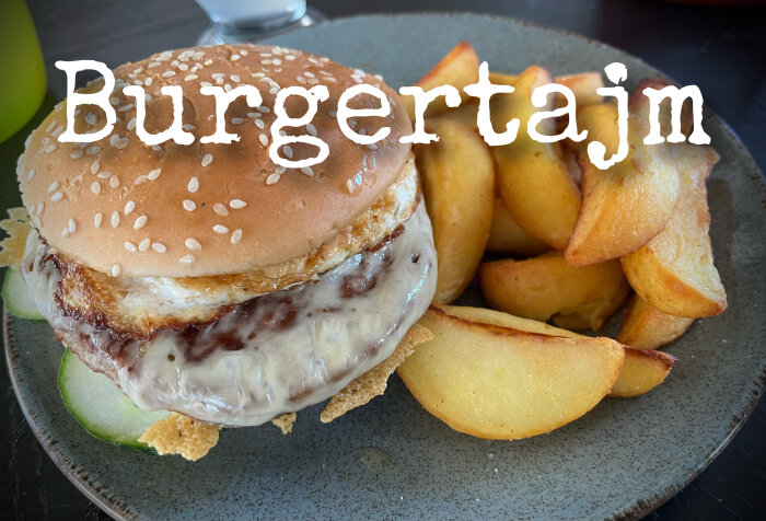 Hamburgare med sesamfröbröd, ost, grönt, sås, och potatisklyftor på en keramiktallrik. Text överlagrad.