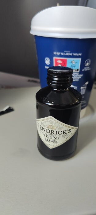 En liten flaska Hendrick's Gin framför en blå mugg på en silverfärgad yta.