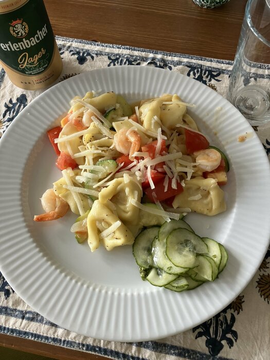 Pasta med räkor, tomater, gurka och ost, ölflaska i bakgrunden, vit tallrik, linne med blommönster.