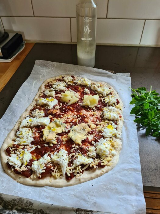 En ougräddad pizza med ost och tomat, olivolja, basilika, på bakplåtspapper på ett köksbänk.