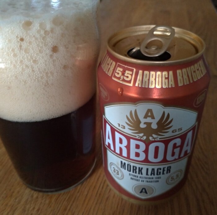 Mörk lager öl i glas och öppnad burk, skumkrona, "Arboga" etikett, dryckesnjutning.