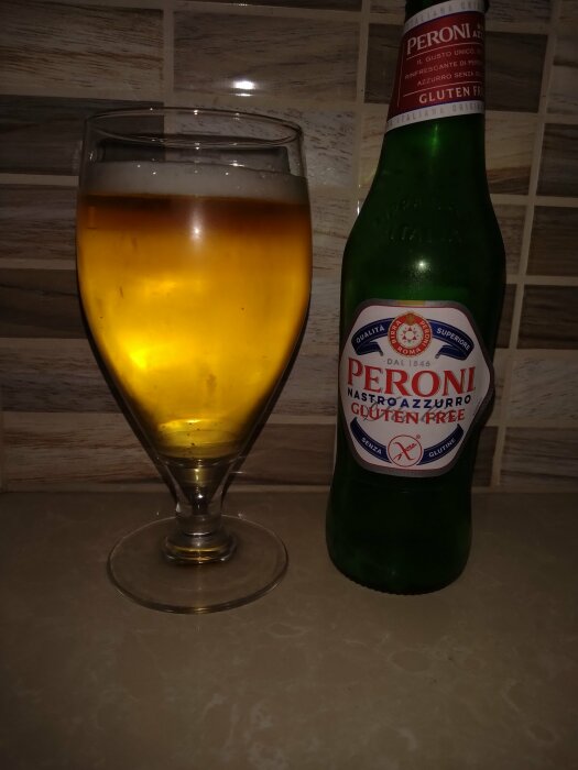 Ett glas med öl och en ofullständigt visad Peroni glutenfri ölflaska, på en köksyta med kakelvägg i bakgrunden.