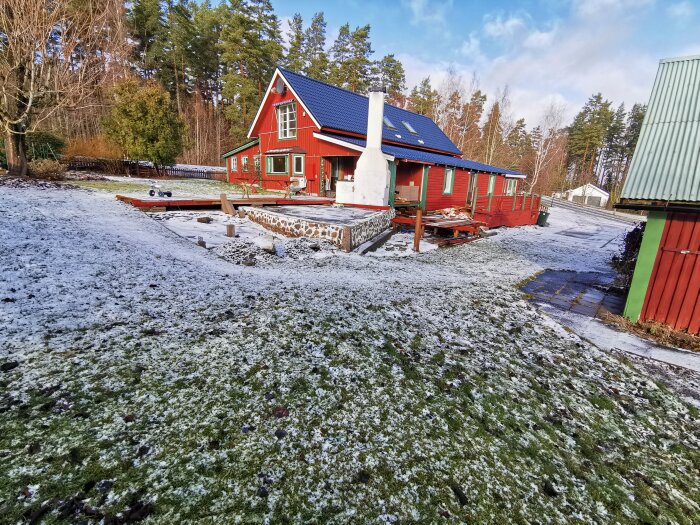 Rött trähus med blått tak, snöfläckar på marken, byggnadsmaterial framför, träd, molnig himmel.