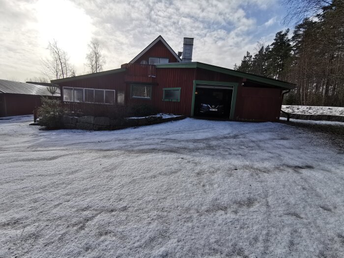 Ett rött hus med garage, snötäckt mark, delvis soligt, skogsbakgrund, stilla vinterscen.