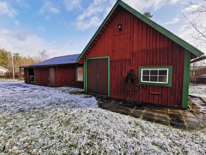 Röd träbyggnad med snöfläckat gräs, soldis, blå himmel, carport, och bar trädgren.