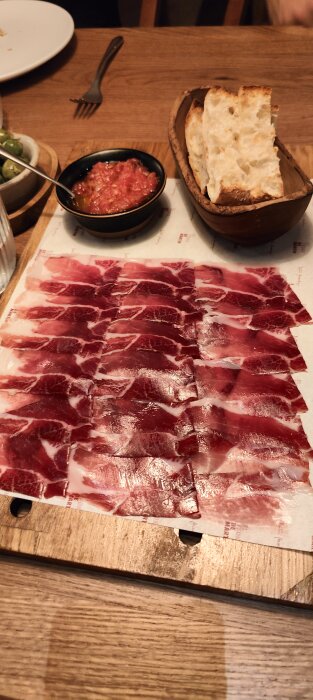 Skivor av lufttorkad skinka framför bröd och tomatsås på träbord. Delikatesser för måltid.