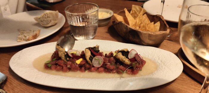 Förrätt med tärnat rått kött, krutonger, rädisor, purjolök, ätbara blommor på vit tallrik; tortillachips, bröd, vin i bakgrunden.