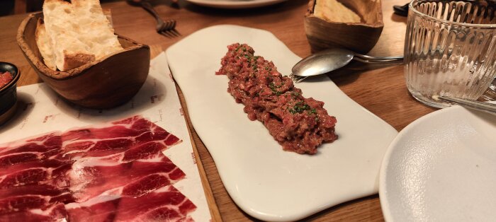 Ett bord med tapas: skivor av rått kött, bröd i träskål, tom glas.