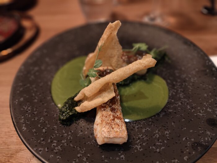 Gourmeträtt med fisk, grönt mos, krispiga chips, mikrogrönsaker på mörk tallrik. Fine dining-presentation.