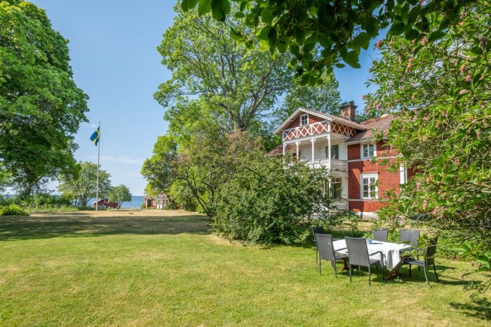 Röd svensk villa, grönskande trädgård, svensk flagga, trädgårdsmöbler, soligt, havsutsikt, sommardag.