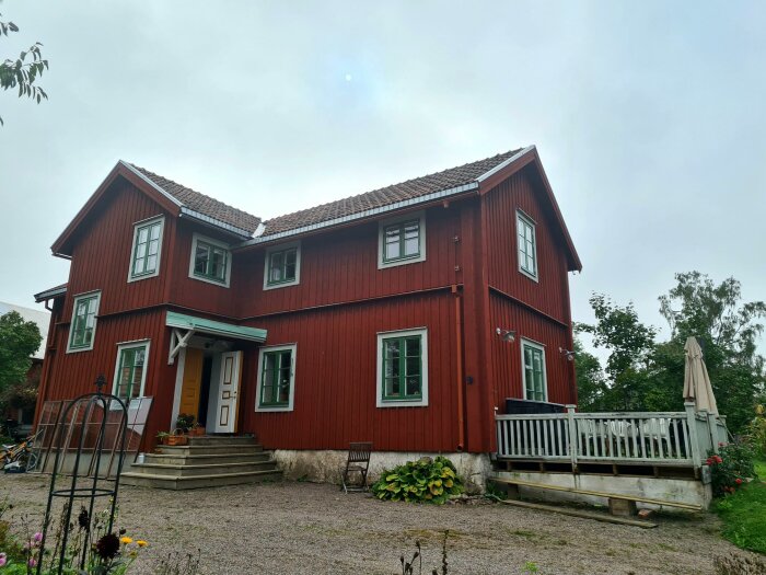 Röd tvåvånings trävilla, vita fönster, altan, trädgård, molnig himmel, klassiskt skandinavisk arkitektur.