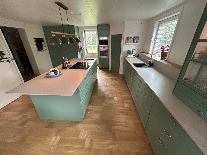 Ett modernt kök med gröna skåp, vitvaror, en ö och trägolv. Fönster ger naturligt ljus.