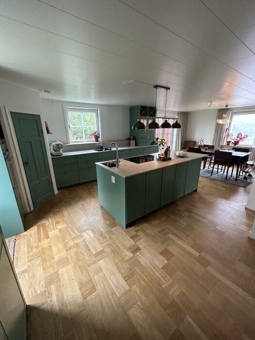 Modernt kök i öppen planlösning med trägolv och gröna skåp, köksö, köksfläkt, fönster och matplats.