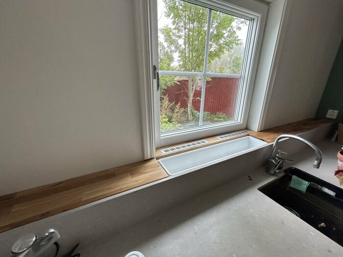 Ett köksfönster med utsikt över trädgården, träbänkskiva, diskbänk, och en kran med diskmedel bredvid.