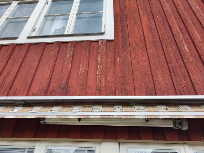 Rödmålad träfasad med slitage, fönster, vita karmar, takränna, del av byggnad.