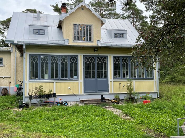 Ett gult hus med vit veranda, fönsterluckor, barnleksaker framför, i en grönskande trädgård.