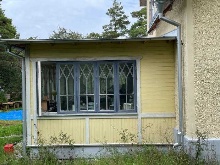 Gul träfasad, vit veranda, fönster med krysspröjs, grönska, dagsljus, friliggande hus, avslappnad miljö.