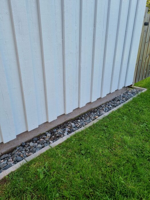 Vit staketvägg, grusremsa, kantsten, grönt gräs. Utemiljö, gräns mellan gräsmatta och staket.