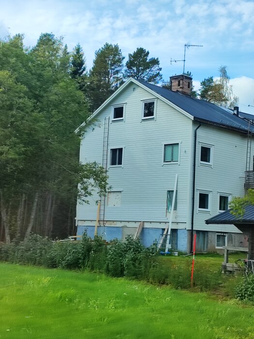 Tvåvåningshus, vit, renovering pågår, grönt gräs, träd bakom, blå himmel med moln, stege vid fasad.