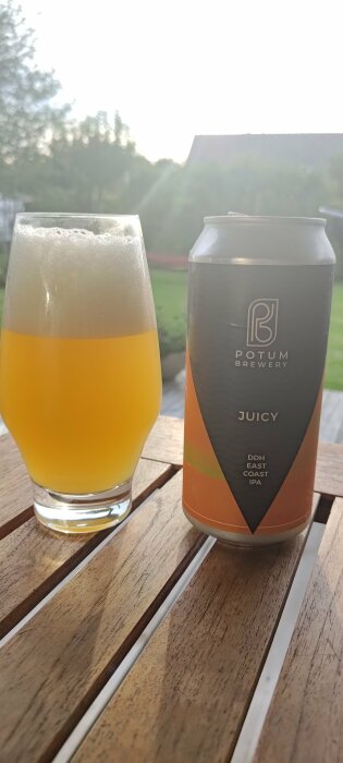 Ölglas på träbord utomhus nära burk med text "POTUM BREWERY JUICY DDH EAST COAST IPA". Solnedgång i bakgrunden.