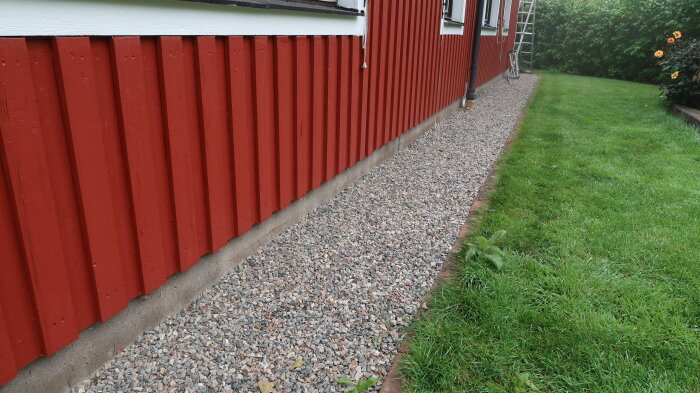 Rödfärgat trähus, grå stenkross längs väggen, grön gräsmatta, stege i bakgrunden, molnig himmel.