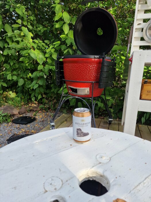 Röd klotgrill öppen, ölburk på träbord, gröna blad, utomhusmiljö.