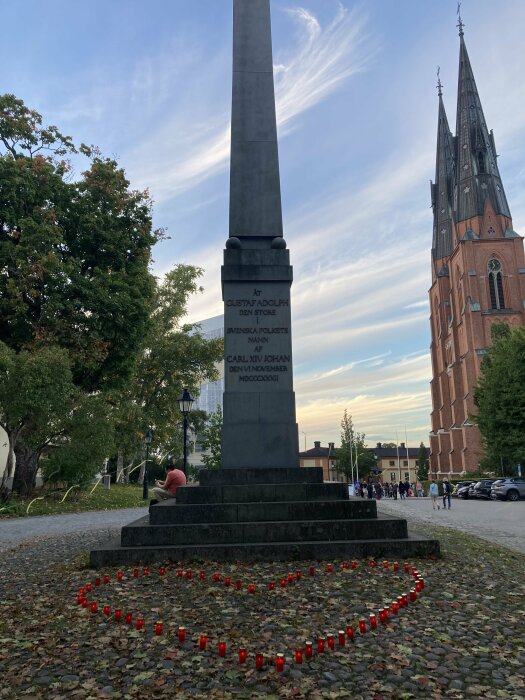 Monument, ljus i hjärtform, kyrka i bakgrunden, människor på avstånd, kvällshimmel, löv på marken.