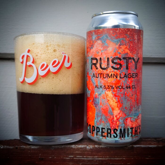 Ett glas mörkt öl med skum bredvid en burk märkt "RUSTY AUTUMN LAGER".