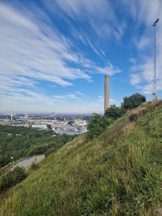 Stor skorsten, kulligt landskap, industriområde i bakgrunden, klar himmel med vita moln, ljusstolpar och gröna buskar.