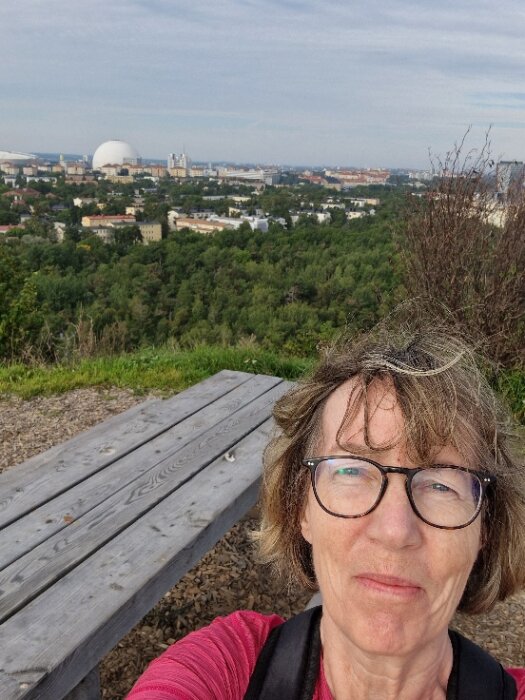 Kvinna tar selfie framför stadssilhuett från utsiktspunkt med träd och parkbänk.