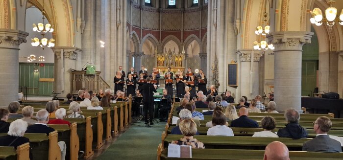 Kör och orkester framför musik för publik i kyrka. Dirigent leder. Ljus, bågar och dekorativa element synliga.