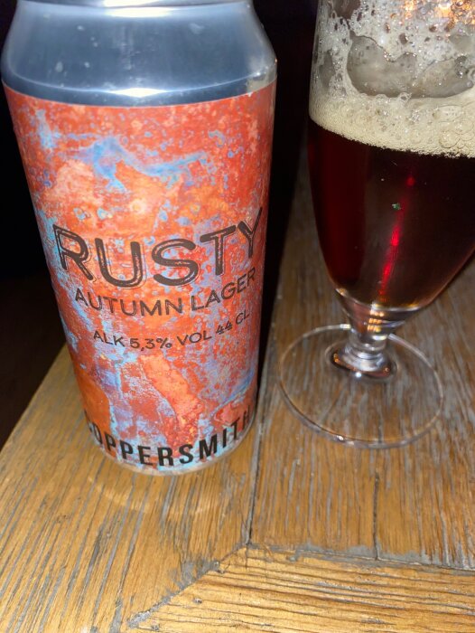 En ölburk märkt "RUSTY AUTUMN LAGER" bredvid ett delvis fyllt ölglas på ett träbord.