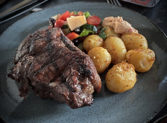 Stekt biff, rostad potatis, sallad, och sås på en mörk tallrik.