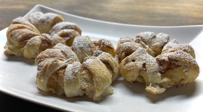 Färska bakverk med florsocker på vit tallrik, ser ut som kanelbullar.