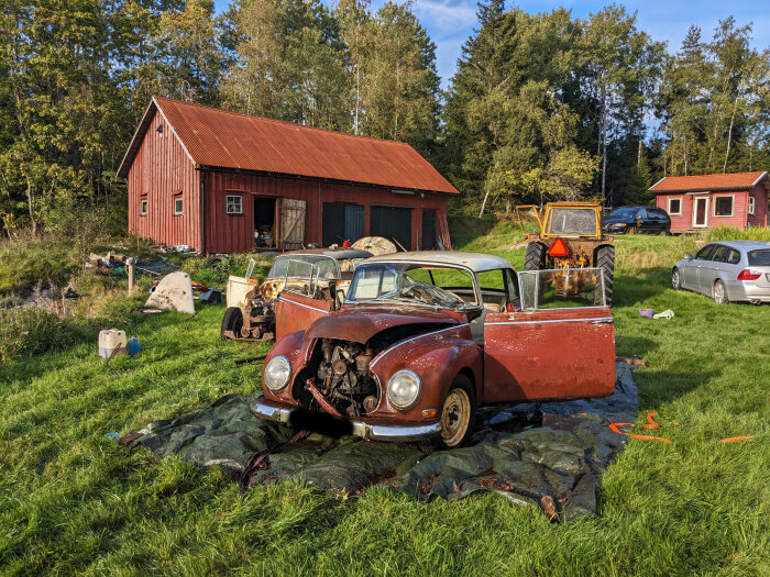 Röd lada, delad gammal bil, traktor, skrot, grönt gräs, skogsbakgrund, klart väder.