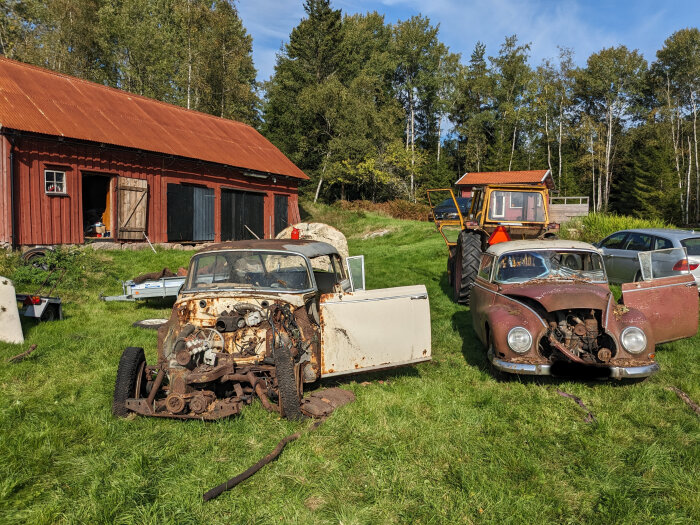 Bild av övergivna, rostiga bilar på en gräsmatta framför röd lada, med traktor och skog i bakgrunden.