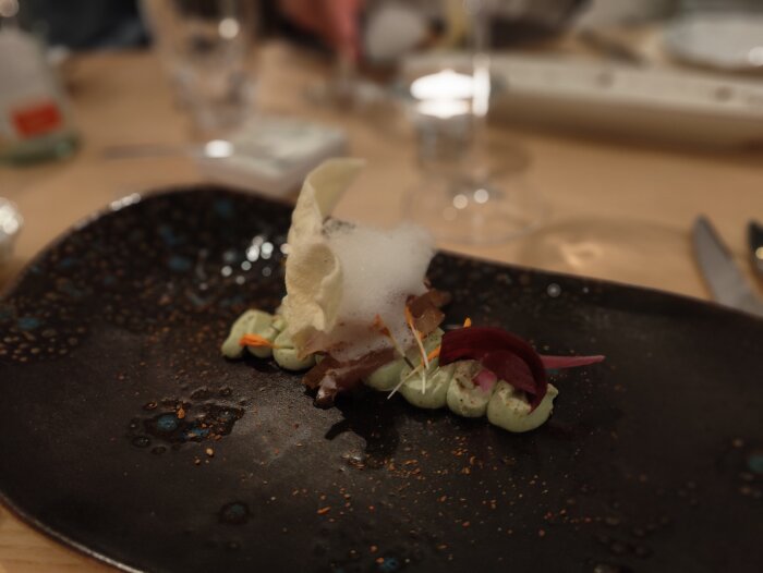 Kreativ maträtt på mörk tallrik, skum, krispiga element, färgglada ackompanjemang, inomhus, restaurangmiljö.