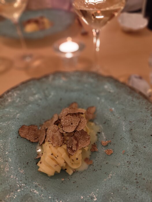 Förrätt med pasta och tryffel på en blå tallrik, ljus bakgrund, suddig vin- eller cocktailglas. Elegant middagsstämning.