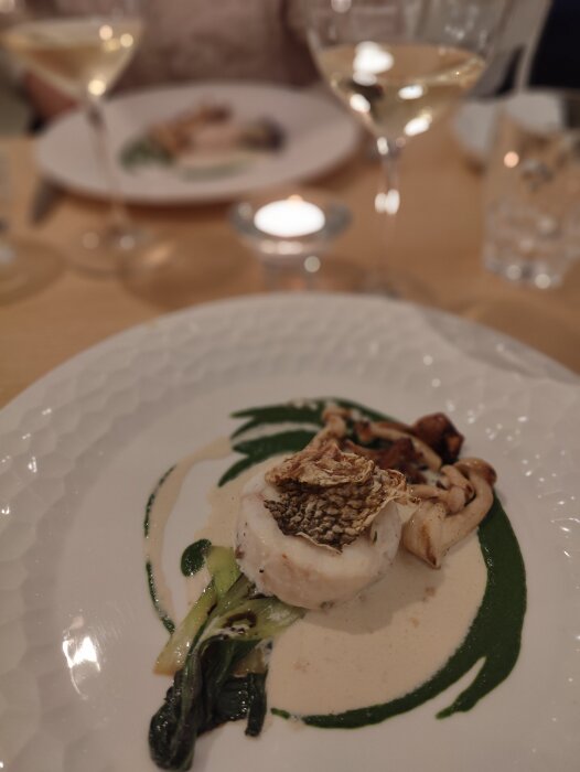 Elegant, upplagd middagstallrik med fisk, sås, grönsaker; i restaurangmiljö med vin, ljus. Fokus på närmaste rätt.